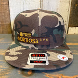 Camo 6-panel snapback hat with embroidered SURF HERMOSA AVE text, displayed on a wooden surface.