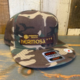 Camo 6-panel snapback hat with SURF HERMOSA AVE patch, displayed on a wooden surface.