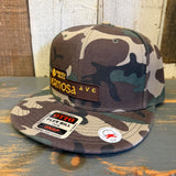 Camo snapback hat with SURF HERMOSA AVE embroidery rests on a wooden surface.