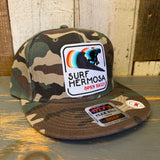 Camo snapback hat with embroidered text on the front, made of fabric, displayed on a wooden surface.