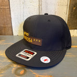 Black 6-panel snapback hat with brown patch, displayed on a wooden surface. Suitable for casual everyday wear.