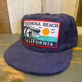 Navy corduroy hat with a patch showing a surfer riding a wave, with birds and sun in the background.