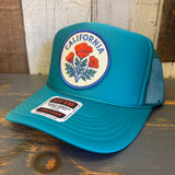 Jade green high crown trucker hat with CALIFORNIA POPPY design, mesh back, displayed on a wood surface.