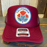 Burgundy high crown trucker hat with poppy design, mesh back, sits on a wooden surface.