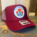 Burgundy maroon high crown trucker hat with mesh back, displayed on a wooden surface.