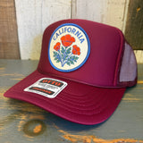 Burgundy high crown trucker hat with California poppy design rests on a wooden surface. Mesh back, casual style.