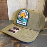 Olive green corduroy cap with 1 to 3 FEET HIGH & RISING patch, resting on a wooden surface.