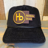 Black corduroy cap with yellow Hb logo, displayed on a wooden surface.