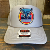 Grey trucker hat with LETS GET SERVED HERMOSA BEACH VOLLEYBALL text, mesh back, displayed on wooden surface.