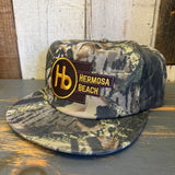 Camo-patterned 5-panel cap in muted tones, made of fabric, rests on a wooden surface. Ideal for casual or outdoor use.
