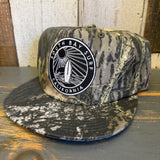 Camo 5-panel surf hat with black patch, displayed on a rustic wooden surface.