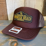 Brown high crown trucker hat with mesh panels rests on a wooden surface.