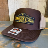 Brown and khaki high crown trucker hat with patch, mesh backing, on a wooden surface.