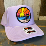 Lavender trucker hat with mesh panels, featuring a sunset, pier, and tower patch on the front.