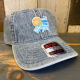 Light blue denim dad hat with Hermosa Beach lifeguard tower design on the front, placed on a wooden surface.