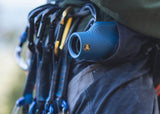 Gray monocular telescope clipped to a climber’s harness with metal carabiners among assorted climbing gear.