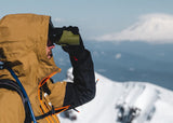 Person in a yellow jacket uses a black NOCS Provisions Zoom Tube 8X on a snowy mountain with rugged peaks behind.