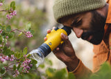 A person in a beanie and orange jacket uses a black monocular to view pink flowers outside.