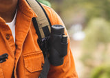 Orange-shirted person outdoors with a black and blue monocular telescope attached to their backpack strap.