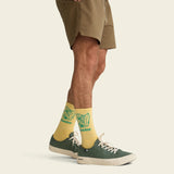 Yellow Howler Brothers El Monito Surfs Socks on a person in tan shorts, paired with green and yellow sneakers, plain background.
