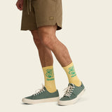 Person in tan shorts and yellow patterned socks with light green sneakers, standing on a plain background.