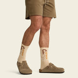 Person wearing beige socks with 'Howler' branding, brown shorts, and shoes on a beige background.