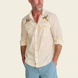 A man wearing a beige shirt and light blue pants on a plain background.