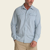 Man in light blue long sleeve UV-protective shirt and beige pants stands against a plain background.