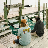 Two people sitting on a dock by water, one wearing a 'Howler Bros' shirt.