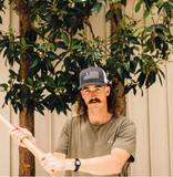 Man in a blue mesh trucker hat and casual wear holds a stick outdoors, with trees and a wall in the background.