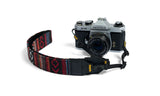 Woven tapestry camera and binoculars strap in multicolor, attached to a vintage film camera on a white background.