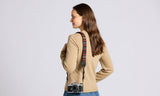 Woman in beige sweater wears a woven tapestry camera and binocular strap, standing and looking to the side.