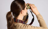 Woman in tan sweater uses binoculars with a woven tapestry strap around her neck.