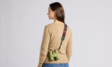 Woman in tan sweater and jeans carries green binoculars with a woven tapestry strap.