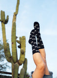 White-patterned crew socks on legs, lifted by a cactus with a blue sky background; cotton blend for casual wear.