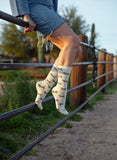Individual wearing denim shorts and patterned crew socks sits on a metal fence with green foliage behind them.