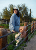 Woman outdoors in denim shorts wearing white cotton crew socks with green stripes, blue sweater, and cap.