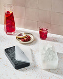 Red drink in glass and pitcher with snacks beside a gray silicone ice tray on a speckled countertop.