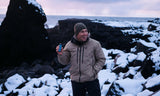 Smiling man in tan jacket on snowy rocks by the ocean holds a compact black and blue 10x25 monocular, ready for viewing.