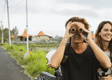 Woman and man hold waterproof black binoculars by a motorbike on a rural road, using them for outdoor viewing.