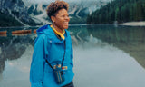 Person in a blue jacket standing by a lake with mountains in the background