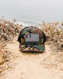 Navy and white mesh trucker hat with a patch, placed on sand by a rocky coastal cliff.