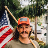 Man in a red mesh trucker hat stands by palm trees, holding a flag. The cap is cotton with a structured front panel.