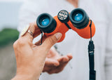Person holding bright orange waterproof binoculars; another pair visible in the background.