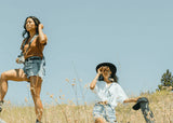 Two women in a grassy field; one takes photos, the other uses green waterproof binoculars with a textured rubber grip.
