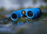 Waterproof binoculars with gold emblem resting on wet rocks beside water. Durable, outdoor-ready design.
