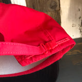 Black and red five-panel cap with mini logo, cotton material, displayed on rustic wooden surface.