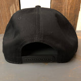 Black snapback hat with classic logo, viewed from the back, resting on a gray wooden surface.