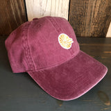 Maroon cotton dad hat with mini logo sits on dark wood in front of a rustic background.