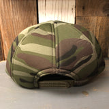 Camo foam winter cap, viewed from the back on a gray surface with wood background.